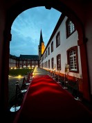 Blick durch einen Torbogen in einen Innenhof mit Rasenfläche. Ein roter Teppich führt aus dem Torbogen heraus. Ein Kirchturm zeichnet sich vor einem Abendhimmel ab. 