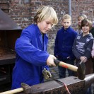 Ein Junge mit Hammer in der Hand bearbeitet ein Stück glühendes Stahlstück auf einem Amboss und eine Gruppe interessierter Kinder schaut ihmm dabei zu.