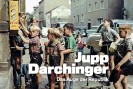 Eine Gruppe jungs in kurzen Hosen stehen an einem kleinen, gelben Bonbonautomaten, der an einer Hauswand befetigt ist.