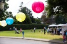 Bunte Lampions gebunden an eine Schnur. Im Hintergrund verschwommen sichtbar Erwachsene und Kinder auf einer Wise, auf der auch Sonnenschirme und ein Stand aufgebaut sind.