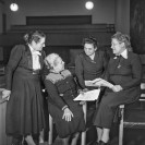 Fotografie von vier älteren Damen in langen schwarzen Kleidern, teils stehend, teils sitzend. Zwei von ihnen halten Zettel in der Hand.