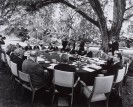 An einem großen runden Tisch sitzen Männer in Anzügen. Der Tisch befindet sich im freien unter einem Baum. Unter den Männern befindet sich der Politiker Willy Brandt.