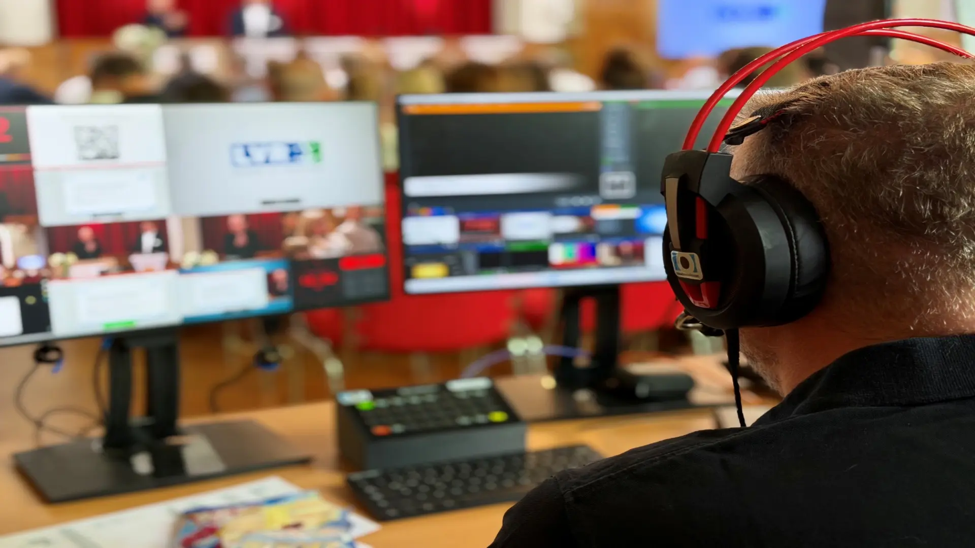 Blick über die Schulter eines Mitarbeiters. Er hat Kopfhörer auf dem Kopf und schaut auf mehrere Monitore. Er sitzt hinten in einem vollen Veranstaltungssaal und kümmert sich um die Technik.
