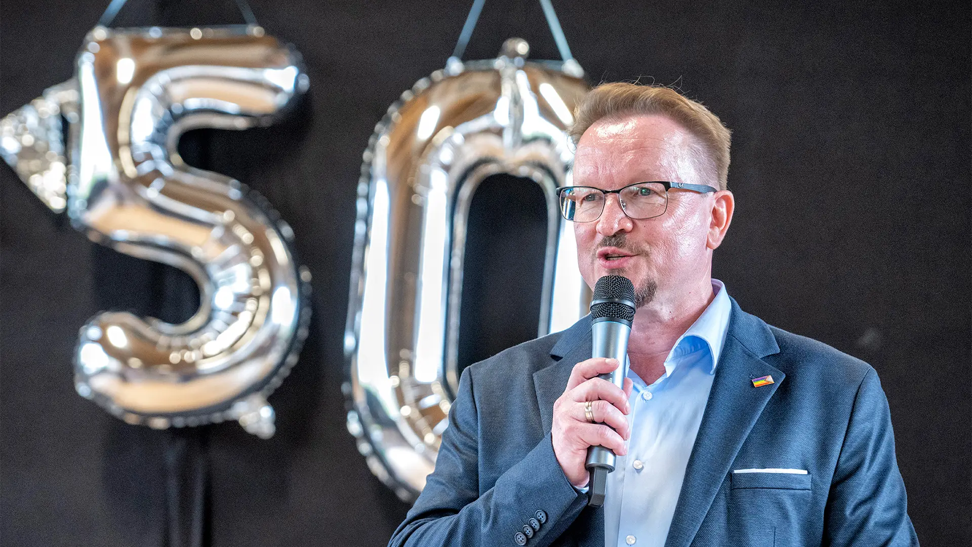 Ein Mann spricht in ein Mikrofon. Hinter ihm zwei Luftballons in Form der Zahl 50