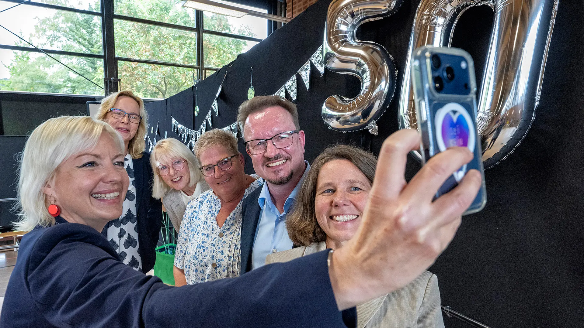 Sechs Personen machen ein Selfie. Neben ihnen an der Wand zwei Luftballons in Form der Zahl 50.