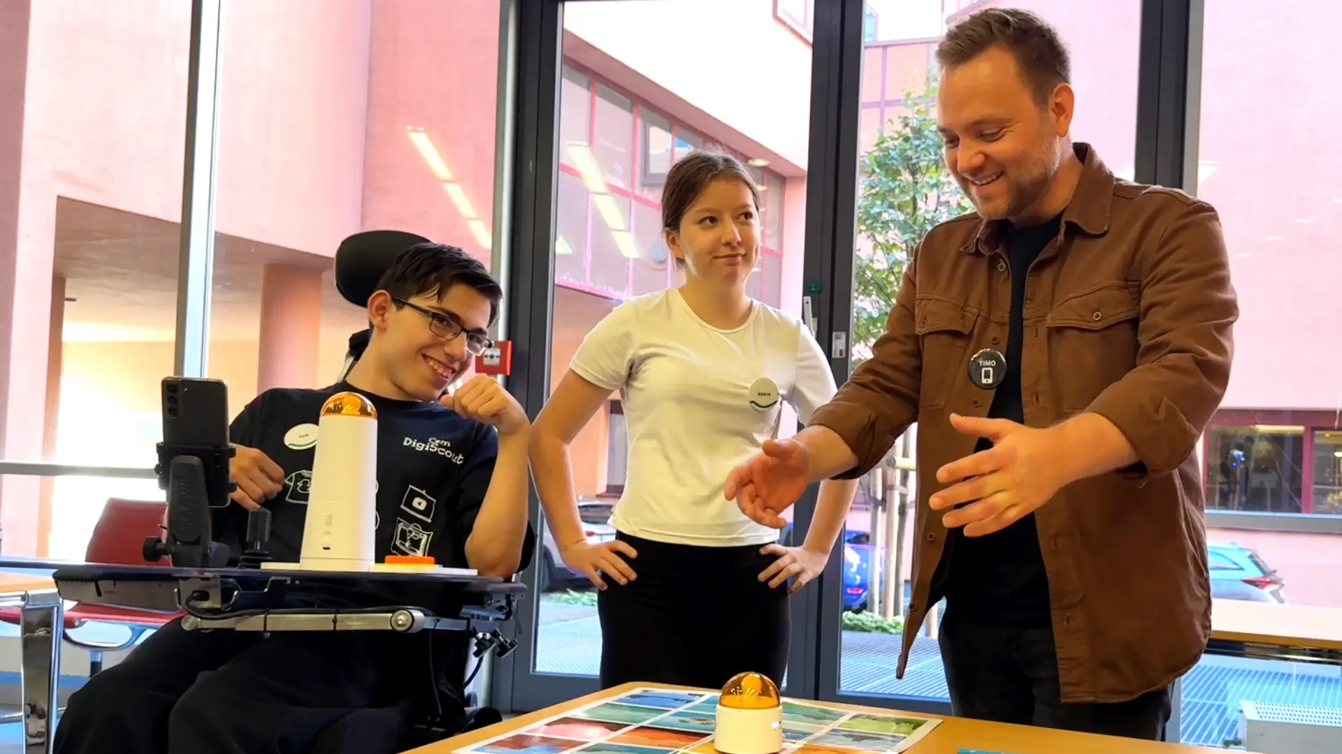 Zwei Schüler*innen und Erwachsener nebeneinander. Der Erwachsene schaut freudig überrascht auf einen Tisch vor ihm. Die Schüler*innen lächeln.