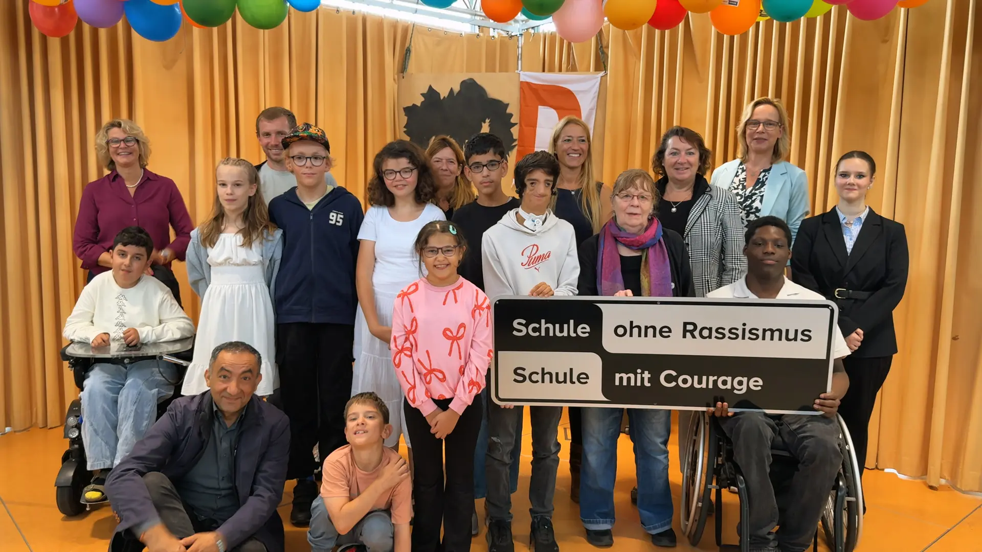 Schüler*innen und Erwachsene auf einer Bühne mit einem großen Schild. Darauf steht "Schule ohne Rassismus - Schule mit Courage".