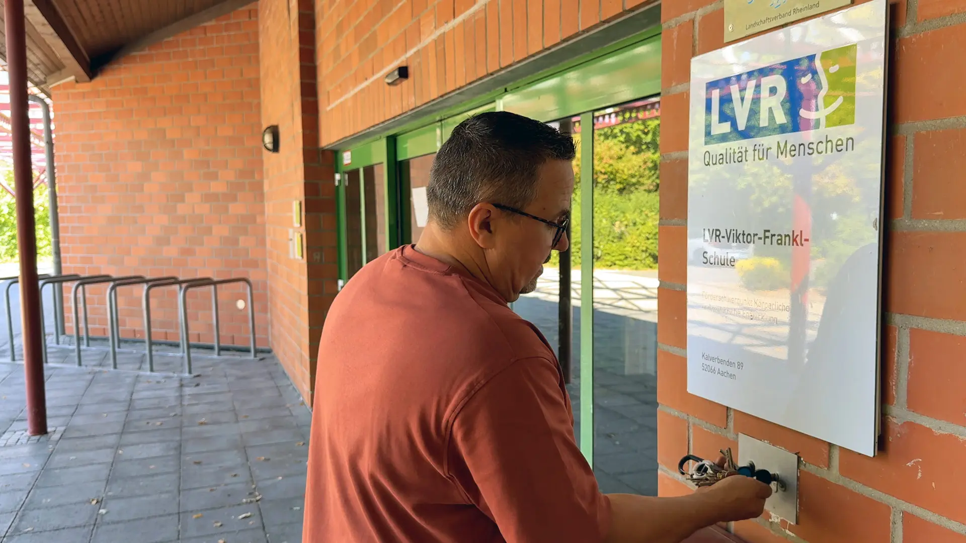 Ein Mann öffnet mit einem Schlüssel dir Eingangstüre der LVR-Viktor-Frankl-Schule. Dabei handelt es sich um eine Schiebetüre.
