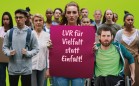 Gruppe aus Menschen verschiedenen Alters und Herkunft, Person in der Mitte der Gruppe hält ein Plakat hoch mit den Worten 