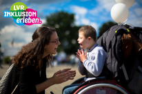 Ein Junge sitzt im Rollstuhl. Vor ihm hockt eine Frau. Beide klatschen in die Hände.