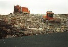 Das Bild zeigt eine Mülldeponie mit zwei orange-farbenen Müllautos