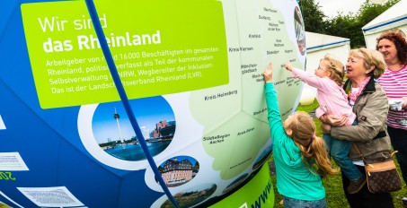 Eine Familie steht vor einer großen Kugel, auf der eine Rheinlandkarte und Bilder aus dem Rheinland zu sehen sind. Sie zeigen auf einen Stadtnamen.