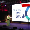 Moderatorin steht auf der Bühne vor einer großen Leinwand, die das 70-Jahre-Landschaftsverbände-Logo zeigt