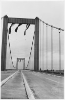Schwarz-Weiß-Foto einer Brücke mit hohen Masten rechts und links aus Sicht eines Autofahrers