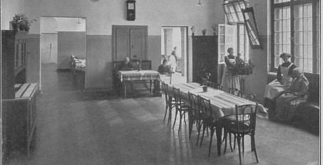 Historische Aufnahme eines Aufenthaltsraums in einem sogenannten Halbruhigenhaus, in der Mitte ein langer Tisch, am Rand stehen und sitzen Menschen.