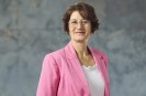 Eine brünette Frau in einem rosa Blazer vor einer grauen Marmorwand.