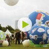 Standbild eines Videos: große blaue und grüne Ballons mit Bildern und Text, zwischen ihnen laufen Menschen