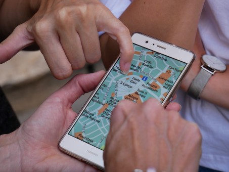 Ein Smartphone mit einer Karte ist im Mittelpunkt des Fotos zu sehen, mehrere Hände gestikulieren drum herum. Die Personen scheinen nach dem Weg zu suchen.