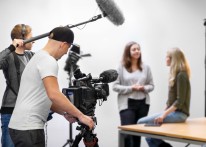 Ein Mann in weißem T-Shirt und schwarzer Mütze steht an einer großen Film-Kamera. Ein weiterer Mann hält ein großes Mikrofon. Im Hintergrund sitzen zwei junge Frauen.