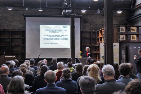 Menschen in einem Saal. Vorn eine Leinwand mit Pr&auml;sentation und eine Person am Rednerpult.