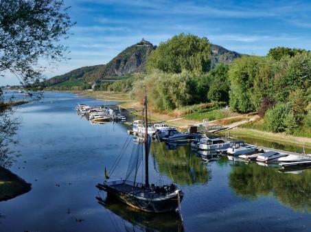 Hier sehen Sie eine Fotografie des Siebengebirges in nordöstliche Richtung. Im Vordergrund fließt der Rhein auf welchem ein Segelboot fährt.