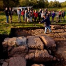 Auf dem Foto ist eine archäologische Ausgrabung zu sehen, bei welcher Baufragmente offengelegt wurden. Im Hintergrund sind mehrere Leute zu sehen, welche den Erläuterungen eines Fachmanns zu dem Ausgrabungsbefund lauschen.