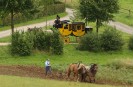 Das Foto zeigt eine historische Postkutsche und einen Bauern, der mit einem Pferdegespann den Boden pflügt