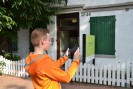 Ein junger Mann mit kurzen Haaren steht vor einem weißen Haus mit grünen Fensterläden und hält ein Tablet in der Hand.