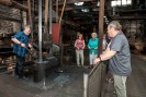 Ein Schmied in einem blaukarierten Hemd bearbeitet ein glühendes Werkstück unter einem Fallhammer. Mehrere Museumsgäste schauen ihm dabei zu.