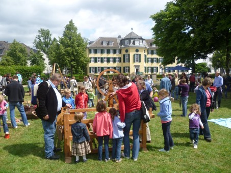 Viele Menschen stehen auf einer grünen Wiese und schauen sich ein nachgebautes Spinnrad an.