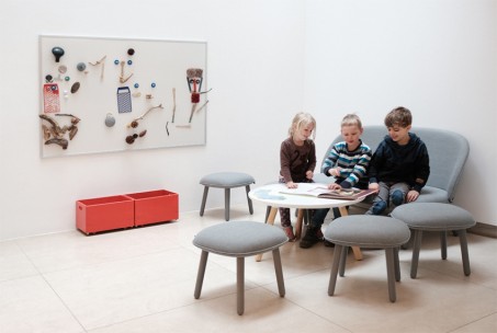 Drei Kinder sitzen auf einem Sofa und blättern in einem Buch, das vor ihnen auf einem Tisch liegt. An der WAnd hängt eine Magnettafel mit vielen Verschiedenen Dingen.