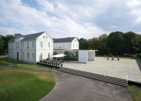 Gebäude-Ensemble mit zwei weißen Häusern und einer großen Terasse aus grauem Stein, eingebettet in eine Parklandschaft