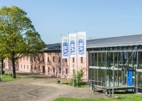 Ein langezogenes Gebäude, dessen Fassade zur linken Seite aus hellrotem Stein, zur rechten Seite aus einer Glas-Stahl-Konstruktion besteht. Vor dem Gebäude stehen ein großer alter Baum und drei Mäste mit Fahnen, auf denen LVR steht.