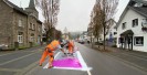 Foto: zwei Männer in orangener Kleidung tragen violette Farbe auf der Straße auf.
