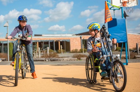 Zwei Schuljungen auf dem Schulhof, der eine sitzt auf einem Fahrrad, der andere ist mit einem Rollstuhl-Fahrrad unterwegs.