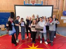 Eine Gruppe von Menschen in einer Sporthalle. Sie freuen sich und halten den Daumen hoch. Vorne zwei Sch&uuml;ler*innen mit dem Schild "Schule ohne Rassismus".