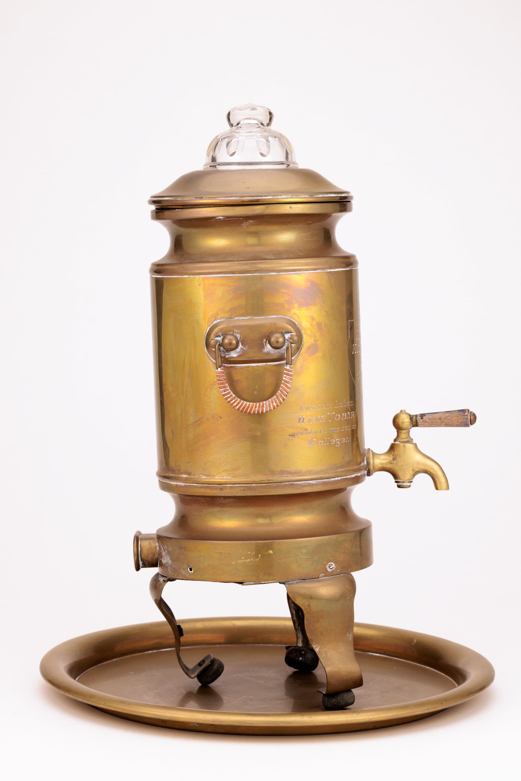 Zu sehen ist ein messing Samovar mit Sicht auf die linke Seite, rechts unten befindet sich ein Hahn, etwa mitting sitzt ein Griff. Der Samovar steht auf 3 Beinen und ist etwas nach links hinten geneigt. Er steht auf einem runden Messing Teller. Der Deckel ist mit einem Glasaufsatz geschmückt.