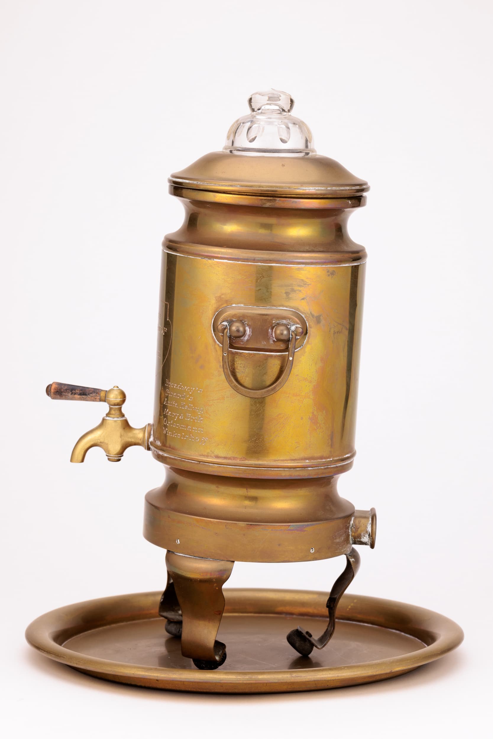 Zu sehen ist ein messing Samovar mit Sicht auf die rechte Seite, links unten befindet sich ein Hahn, etwa zentral sitzt ein Griff. Der Samovar steht auf 3 Beinen und ist etwas nach rechts hinten geneigt. Er steht auf einem runden Messing Teller. Der Deckel ist mit einem Glasaufsatz geschmückt.