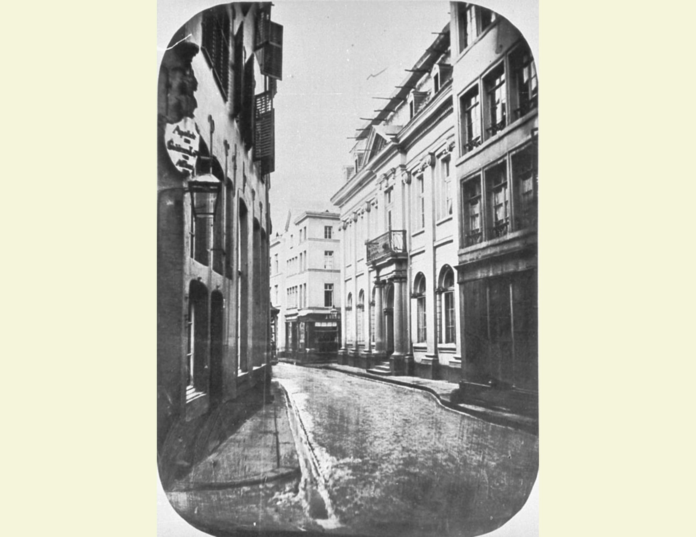 Schwarz-Weiß-Aufnahme einer engen, gepflasterten Straße in einem historischen Stadtviertel. Die Gebäude auf beiden Seiten der Straße sind mehrstöckig, mit sichtbaren Fensterläden und Fassadendetails, die auf eine ältere Architektur hinweisen. Die Straße führt in eine leichte Kurve und verschwindet im Hintergrund des Bildes. Es gibt keine Personen oder Fahrzeuge, was der Schildergasse. Links Antonsgasse mit der Apotheke Zum goldenen Kopf. Rechts Haus Nr. 84 Zum alten Präsidium, Szene eine verlassene Atmosphäre verleiht. Stromleitungen kreuzen den oberen Teil des Bildes und verstärken das historische Gefühl der Aufnahme.