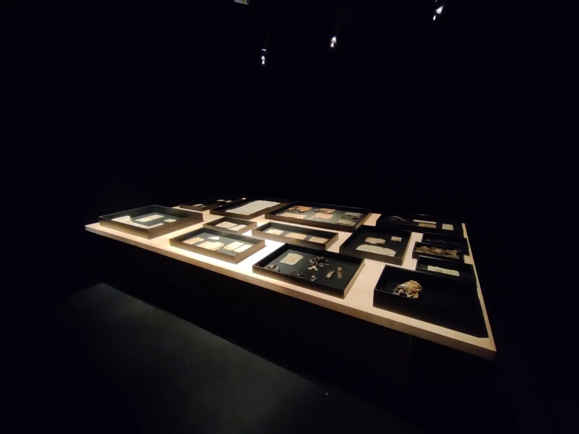 Ein dunkler Raum ist zu sehen. Im Zentrum steht ein Tisch, der mit Licht angeleuchtet wird. Darauf sind verschieden Schriftstücke zu sehen.