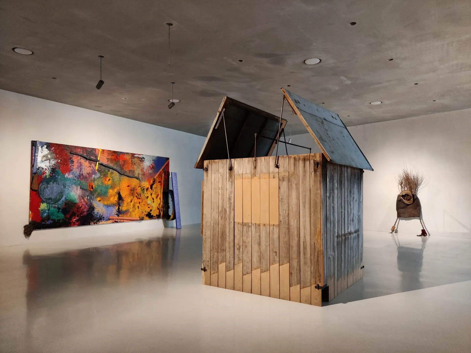 In einem Raum ist eine Laubhütte zu sehen.  An der linken Wand hängt ein buntes Gemälde, an der rechten Seite ist eine Statue zu sehen.
