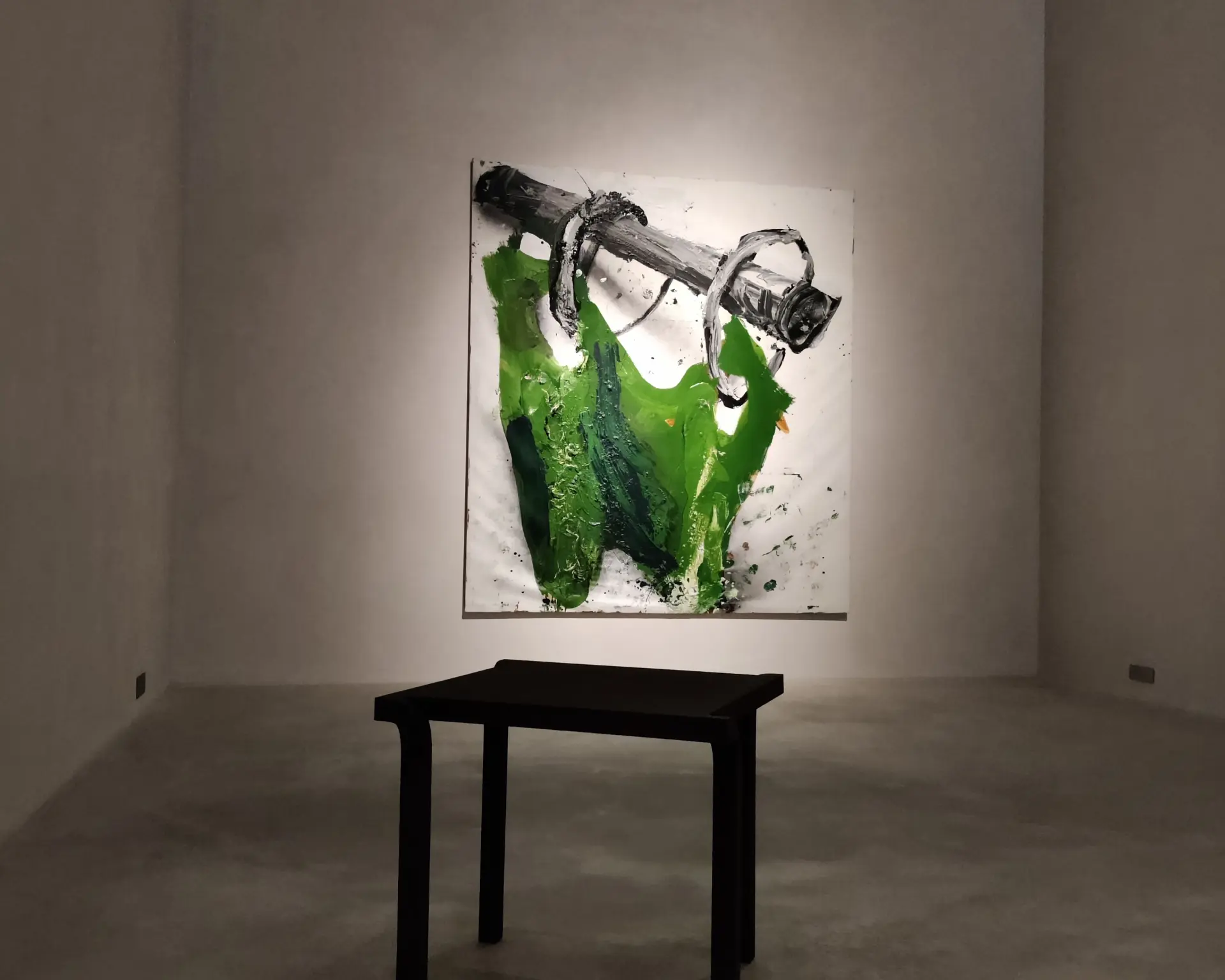 An einer weitßen Wand hängt ein abstraktes Kunstwerk in grün und schwarz, davor steht ein Tisch.