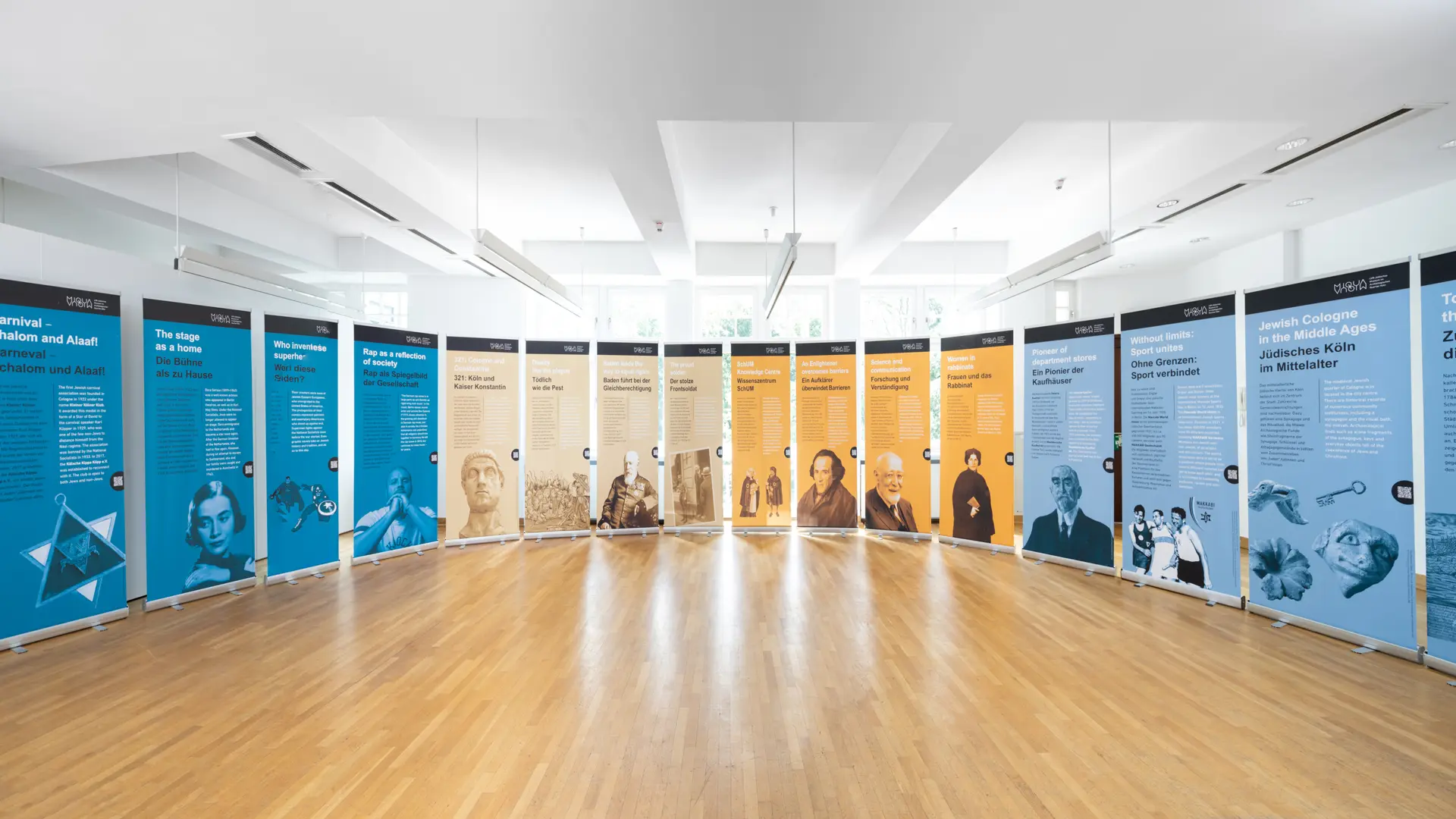 Das Bild zeigt eine Ausstellung mit thematisch gestalteten Informationsroll-ups, die in einem hellen Raum mit großen Fenstern präsentiert werden. Die Roll-ups sind in einem Halbkreis angeordnet und jeweils in den Farben Blau, Orange und Beige gestaltet. Auf den Tafeln sind Titel und Texte in deutscher und englischer Sprache zu sehen, begleitet von historischen Fotografien und Illustrationen. Die Themen reichen von "Jüdisches Köln im Mittelalter" über "Sport verbindet" bis hin zu "Wissenschaft und Forschung". Der Raum hat einen hellen Holzfußboden und weiße Deckenbeleuchtung, die die Exponate gut ausleuchtet. Die große Fensterfront im Hintergrund sorgt für viel Tageslicht und verleiht der Ausstellung eine offene und einladende Atmosphäre. Die Ausstellung ist auf die jüdische Geschichte und deren kulturelle Beiträge in verschiedenen Bereichen hinzuweisen.