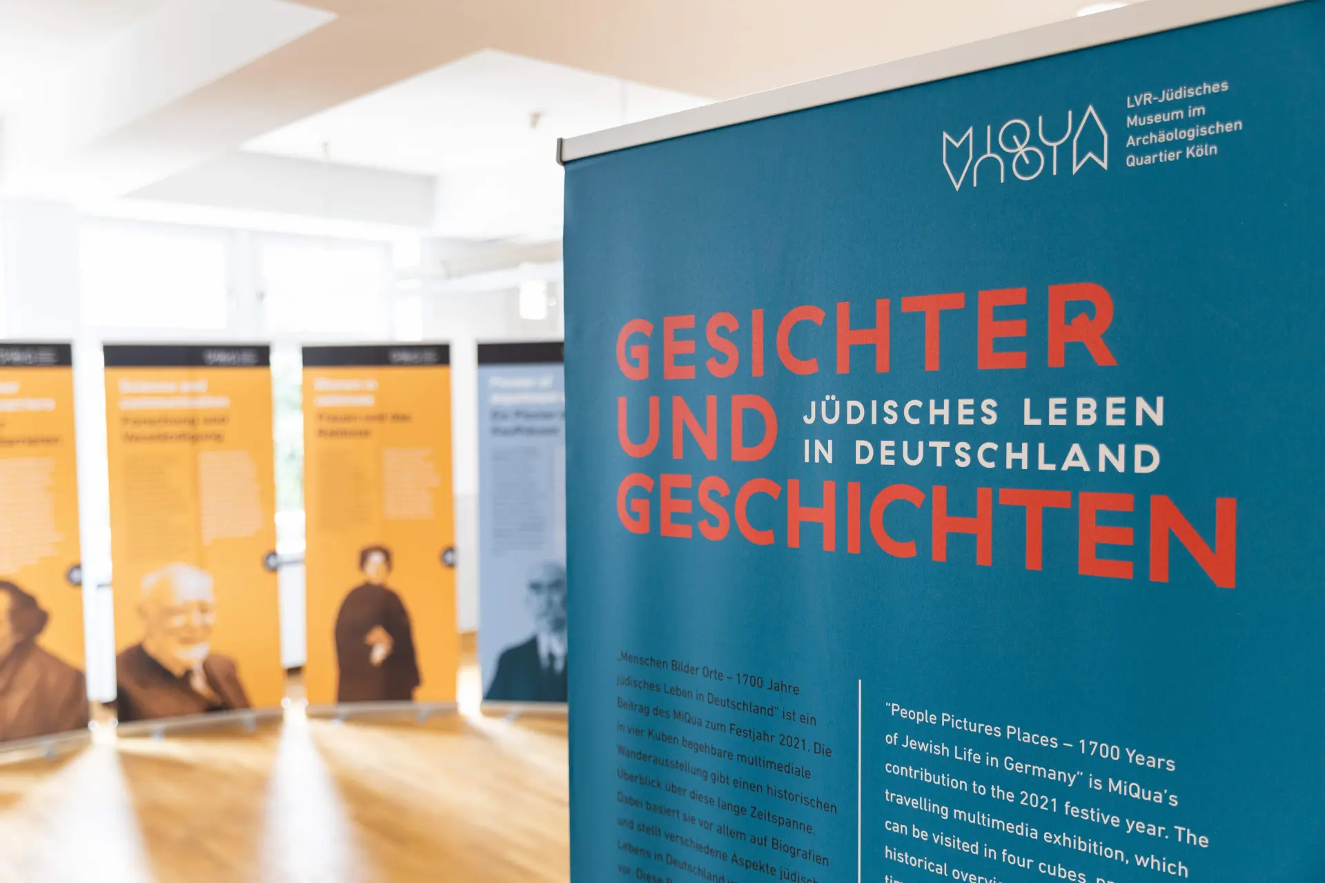 Die Posterausstellung ist in einem hellen Raum mit Holzboden im Zentrum für verfolgte Künste aufgestellt. Im Vordergrund ist eine Nahaufnahme des einführenden blauen Roll-ups zu sehen. Im Hintergrund sind vier Plakate unscharf fotografiert.