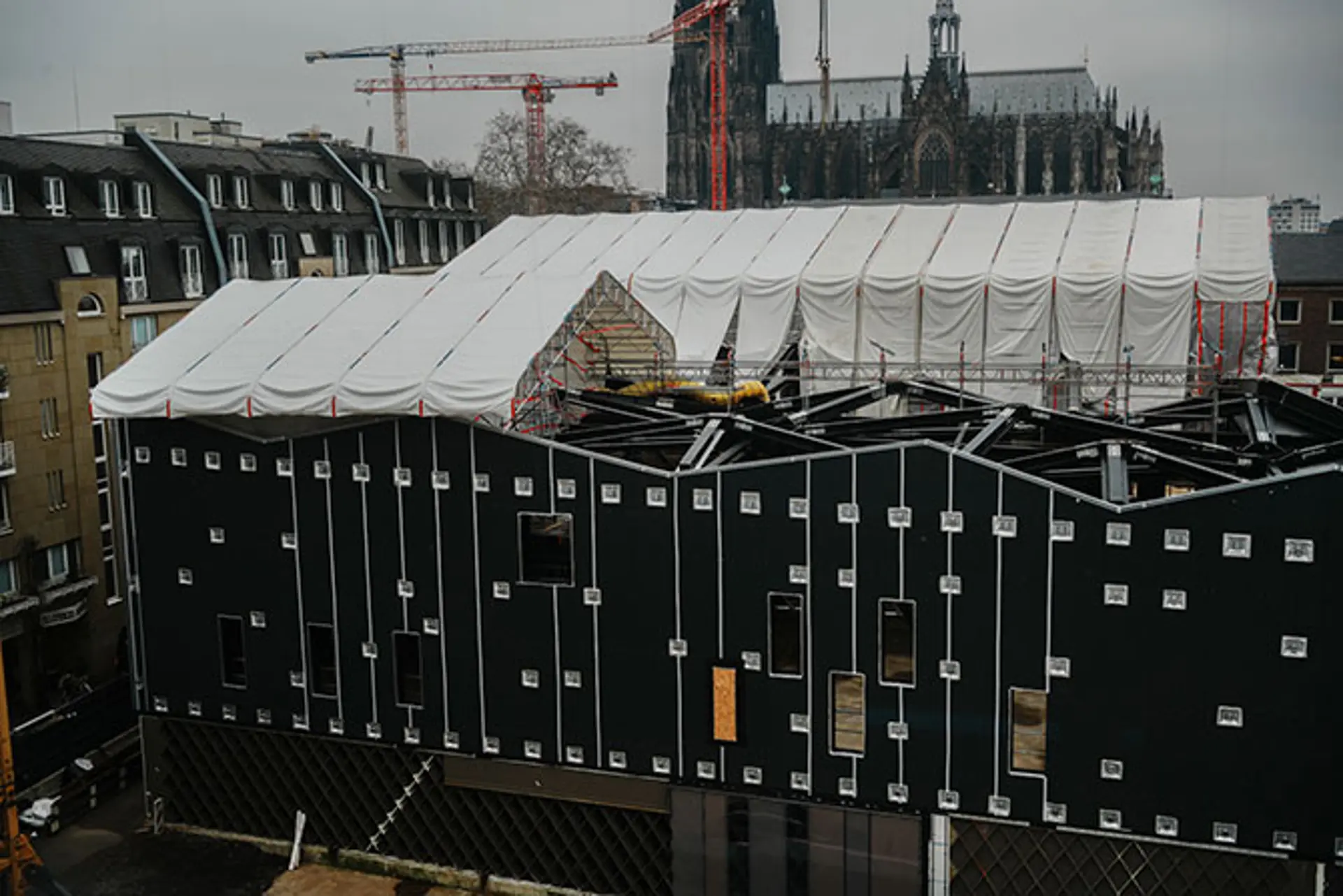 Eine dunkles, zweistöckiges Gebäude ist zu sehen, dass noch im Bau ist.  Ein weißer Bauzaun steht um die komplette Baustelle.  Das Dach ist mit einer weißen Plastikfolie bedeckt.