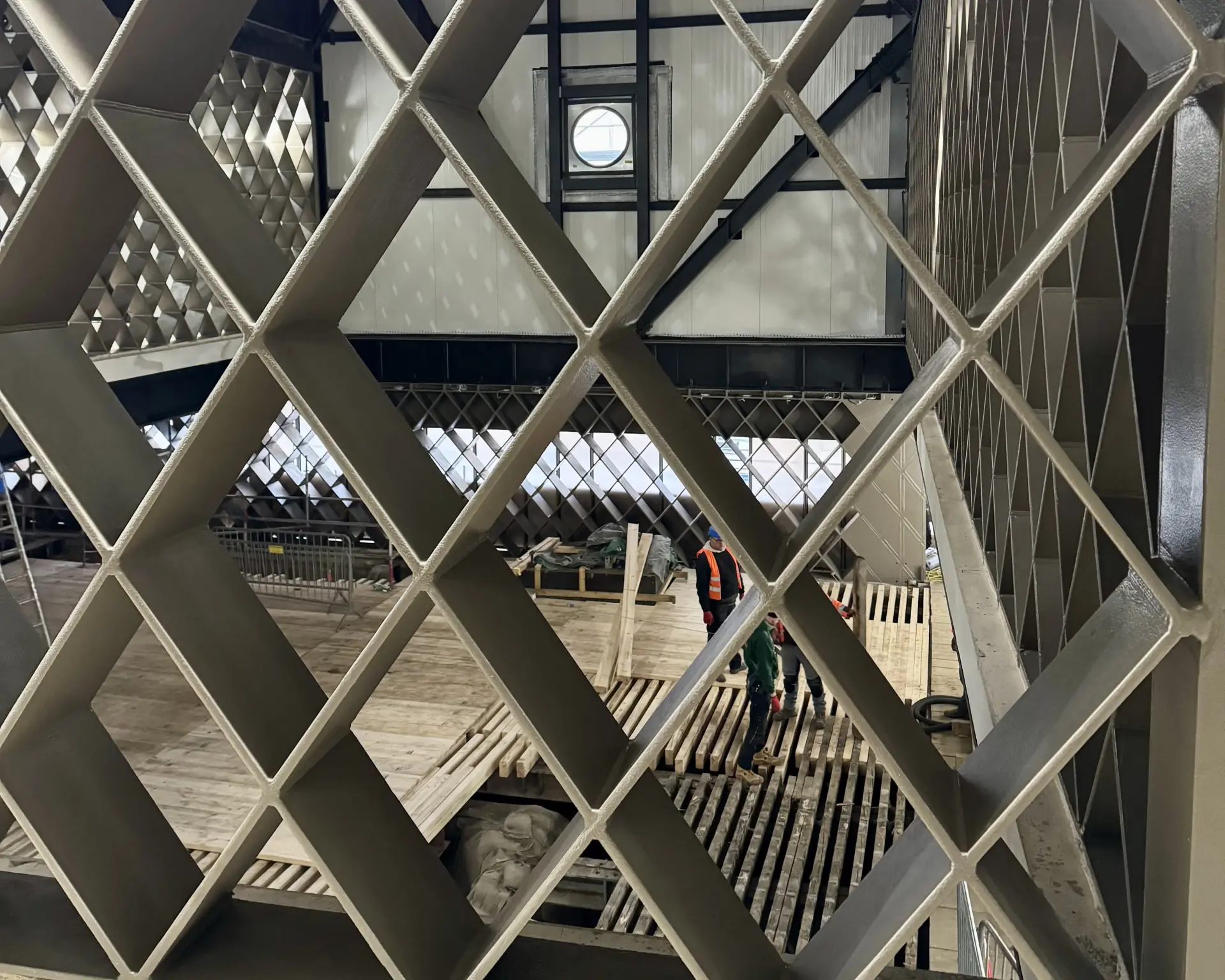 Durch ein Rautenkonstrukt aus Stahl blickt man Auf einer Baustelle blickt man vom Obergeschoss auf einen großen, offenen Raum im Erdgeschoss