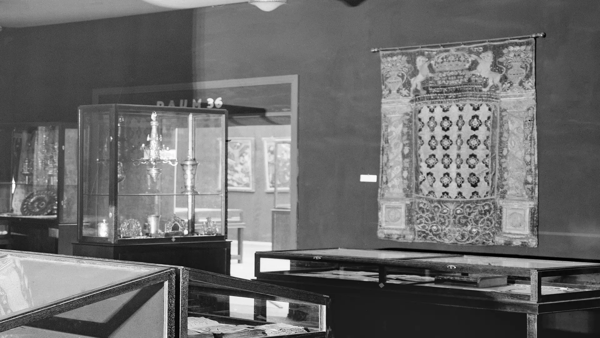 Schwarz-weiß Foto eines Ausschnitts der Jahrtausend-Ausstellung. Der Raum zeigt mehrere Vitrinen, in denen Judaica Objekte ausgestellt werden. An der Wand hängt ein Tora-Vorhang.