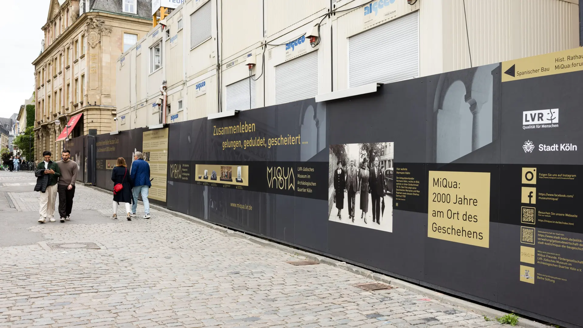 Ein Bauzaun in schwarz und gold rund um eine Baustelle.  Auf dem Bauzaun sind historische Objekte und Ausgrabungsfunde zu sehen.