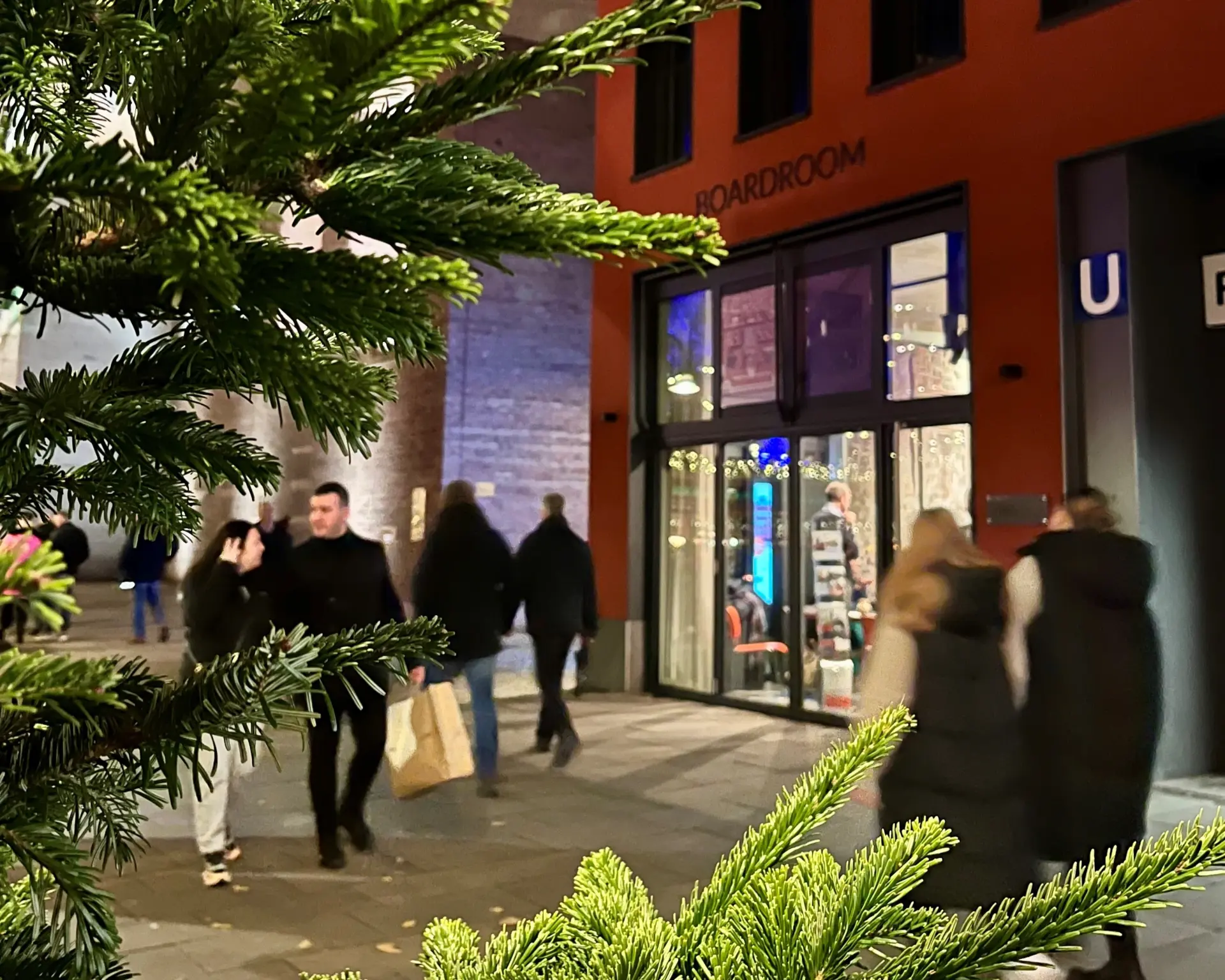 Eine hell erleuchtete Schaufensterfront in einem roten Gebäude.  Links im Bild steht ein Weihnachtsbaum.