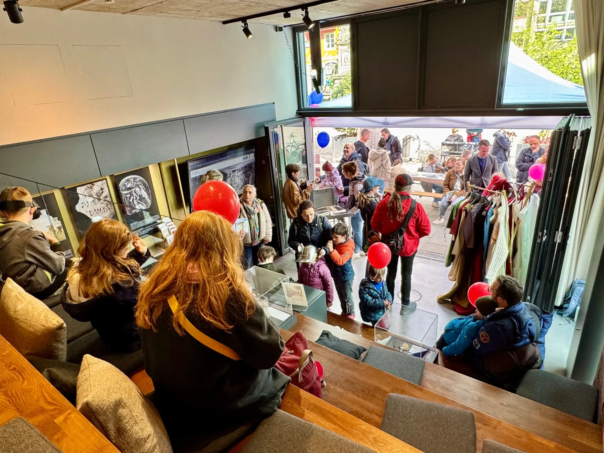 Ein Mann steht vor einem schwarzen Bauzaun und zeigt auf die Abbildung darauf.  Dort ist ein mittelalterlicher Kellerraum zu sehen.  Vor dem Bauzaun steht eine Gruppe Menschen, die dem Mann zuhören.